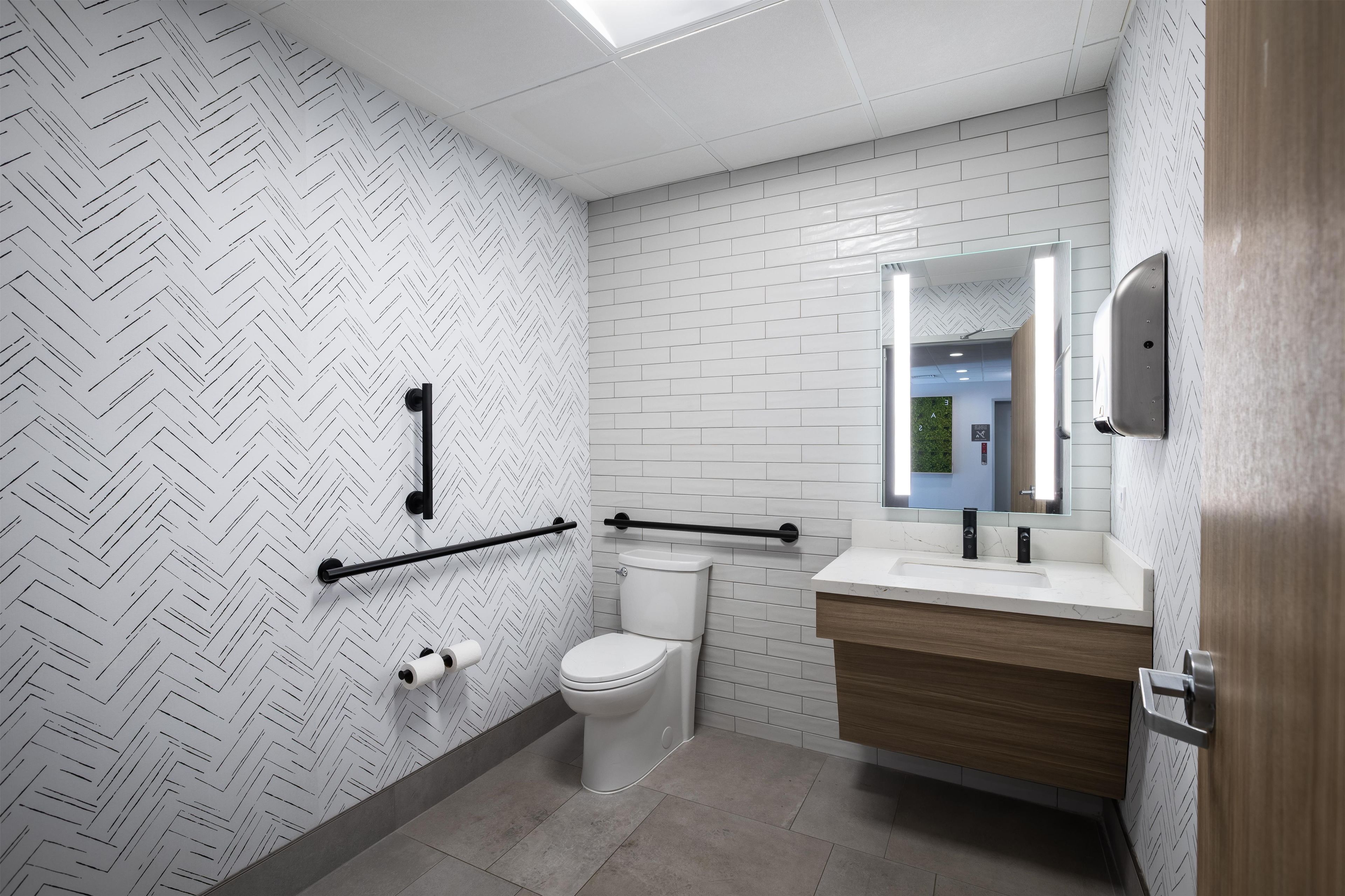 A modern bathroom with a white herringbone patterned wall, a white toilet, a wooden vanity with a mirror, and a black grab bar.