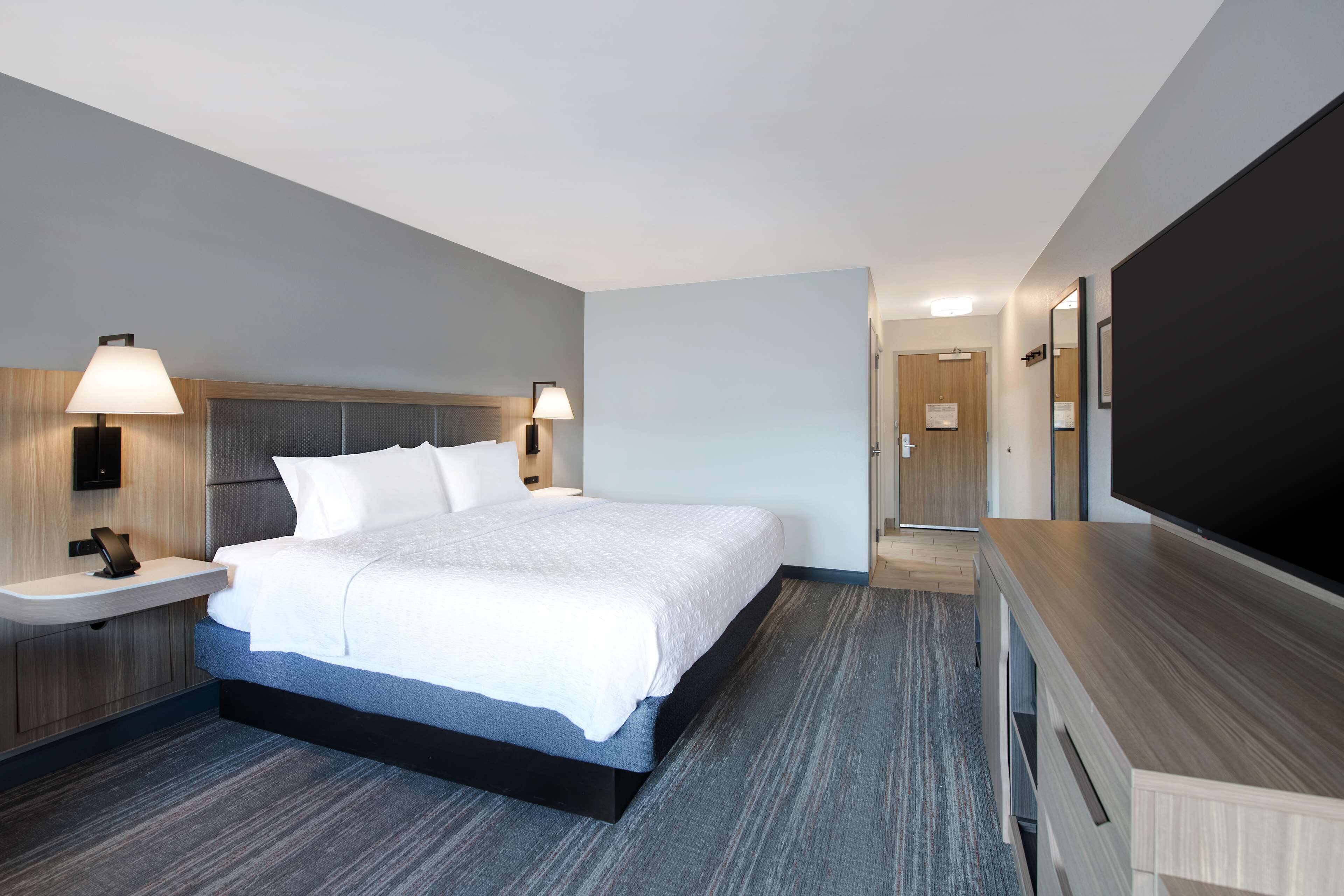 A modern hotel room with a large bed, bedside lamps, a flat-screen TV, a wooden nightstand, and a built-in desk.