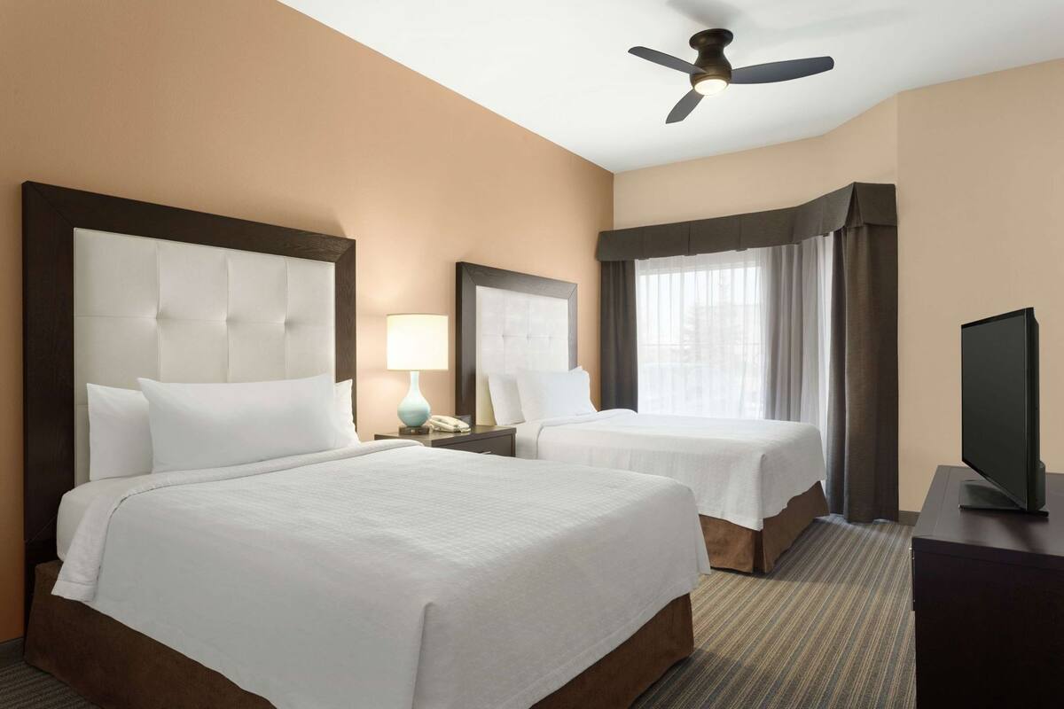 A hotel room with two beds, a ceiling fan, a TV, and a window with curtains.