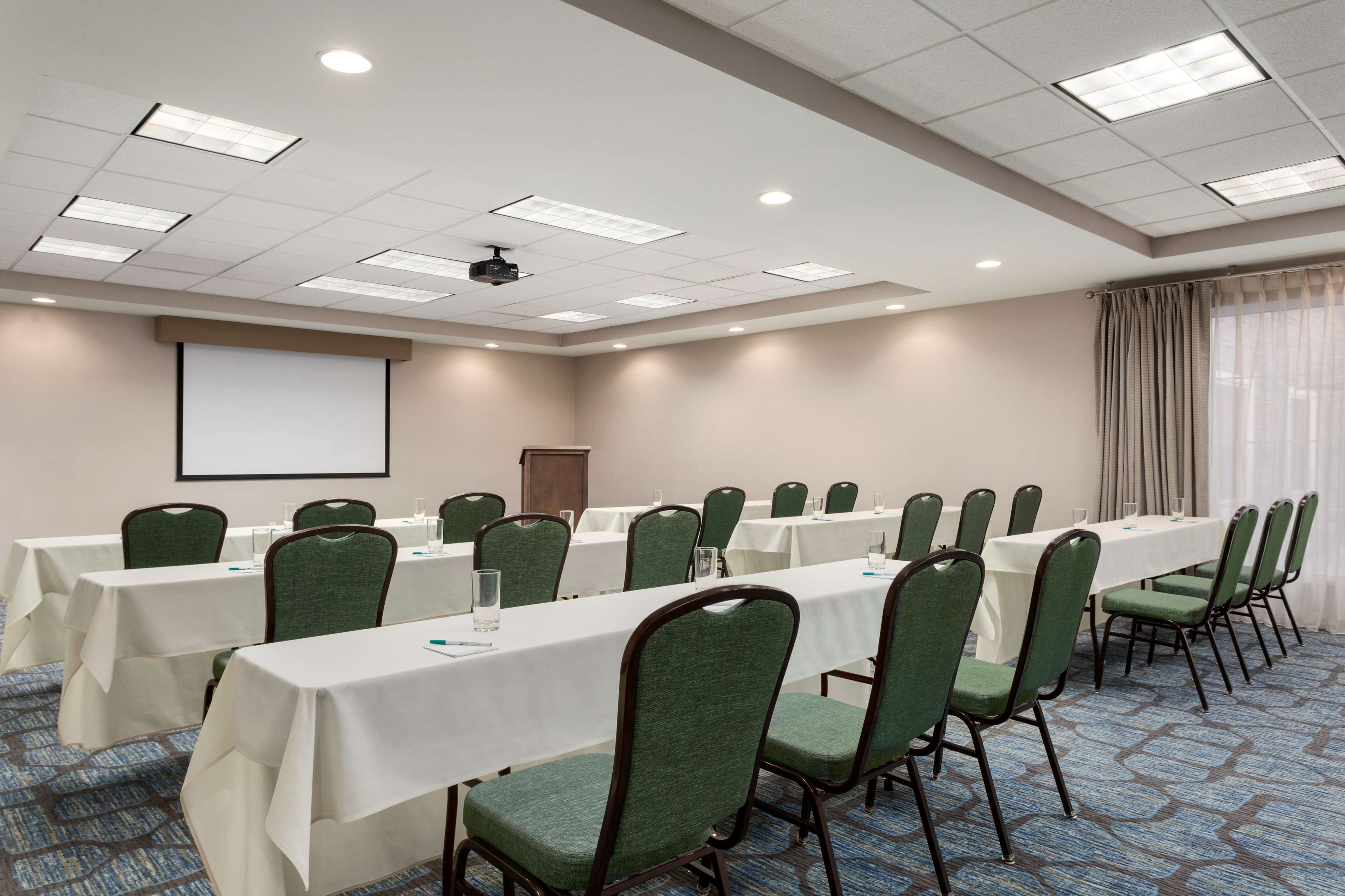 A conference room with long tables, green chairs, a projector screen, and a whiteboard.