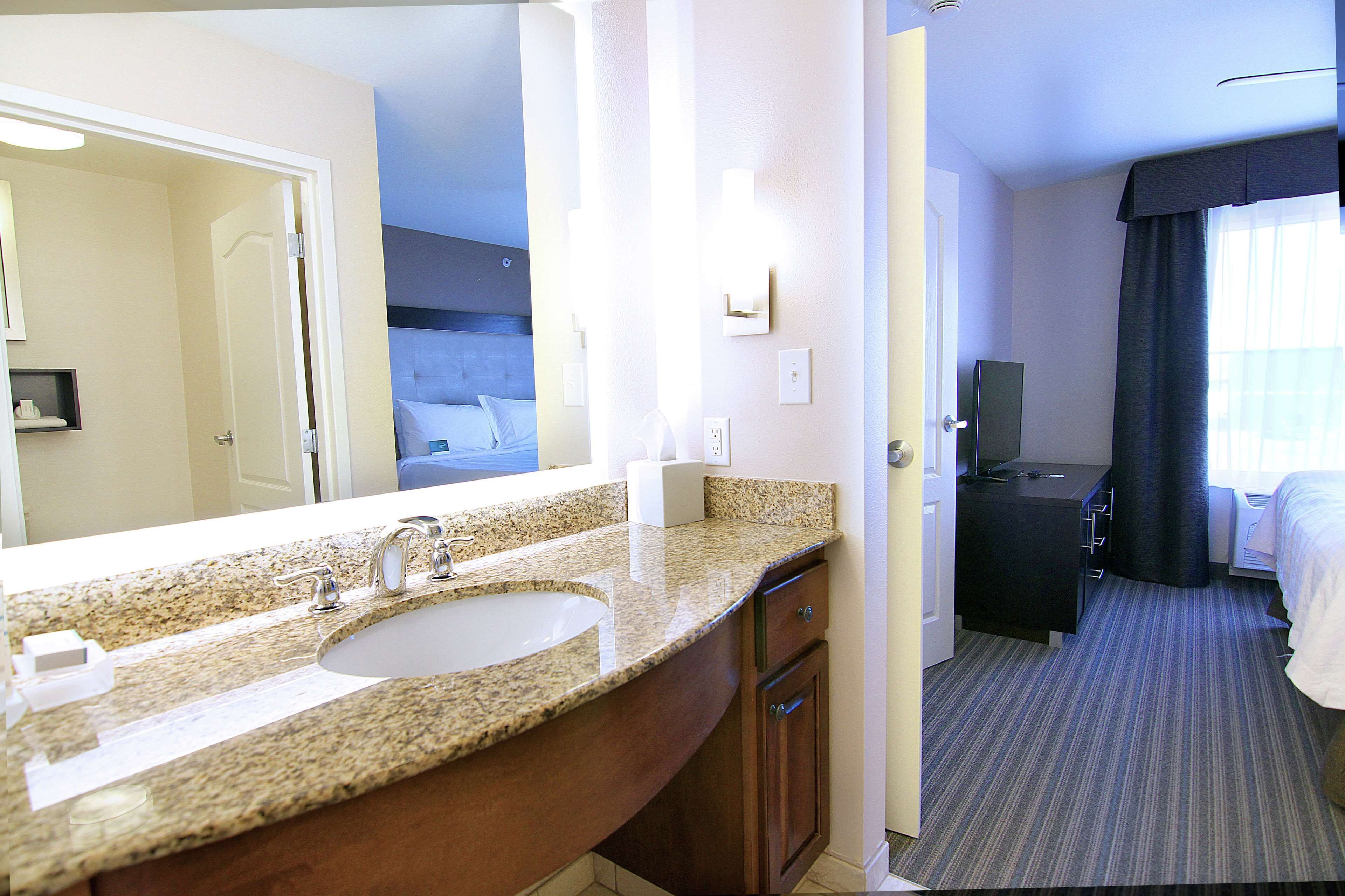 A hotel bathroom with a granite countertop sink, a mirror, and a vanity with drawers.