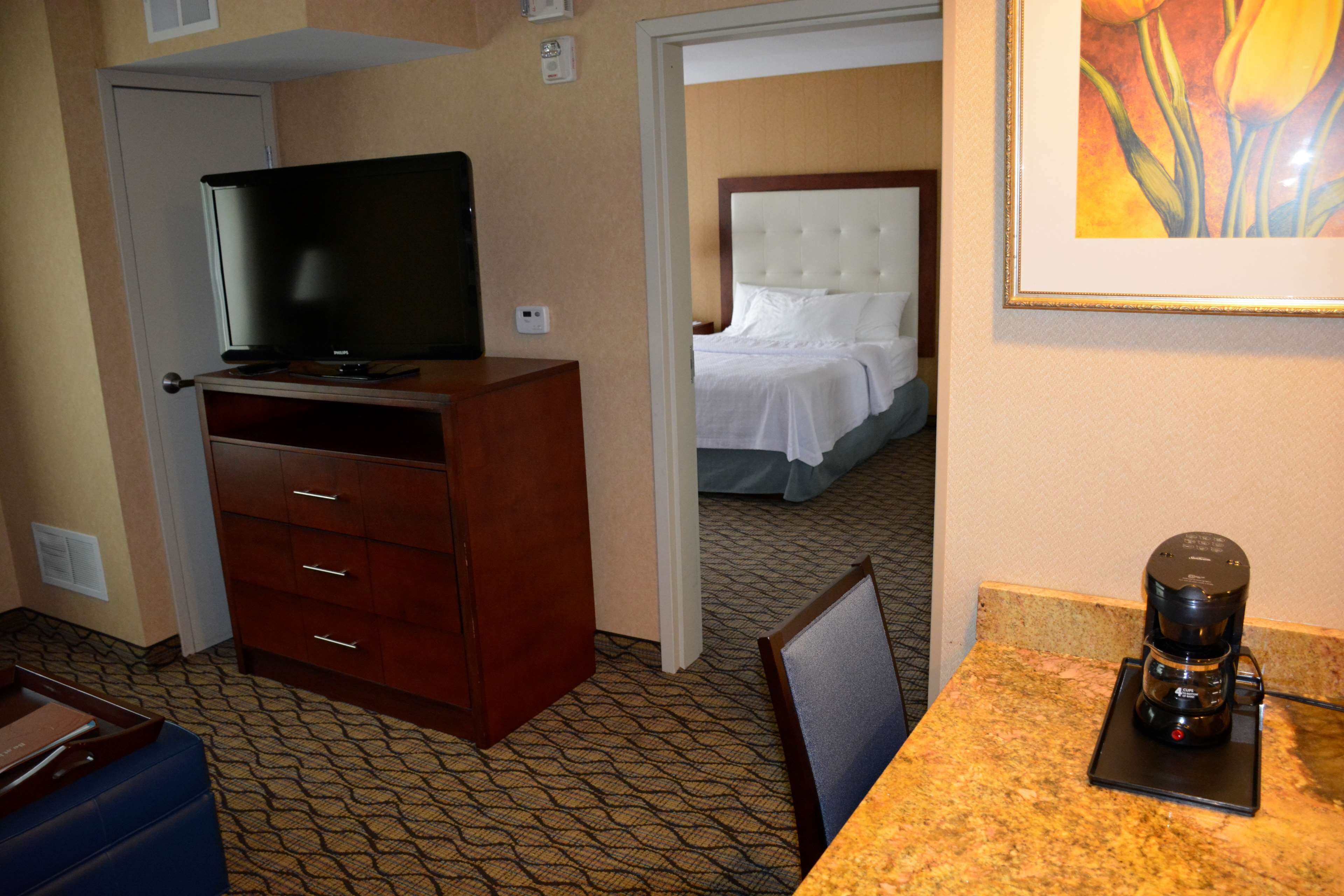 Habitación de hotel con televisión sobre un mueble de madera, cafetera sobre una encimera de mármol y cama con ropa blanca al fondo.