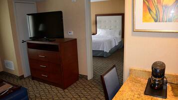 Habitación de hotel con televisión sobre un mueble de madera, cafetera sobre una encimera de mármol y cama con ropa blanca al fondo.