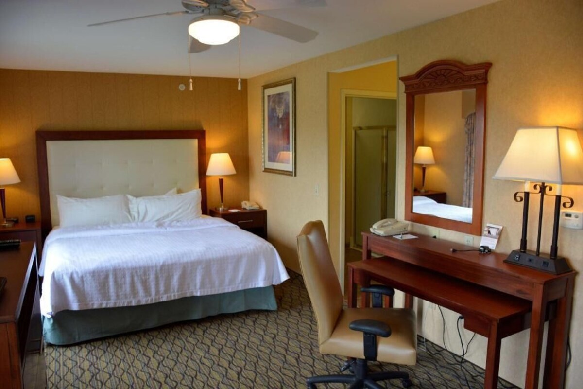 A hotel room with a bed, desk, chair, desk lamp, telephone, and a ceiling fan.