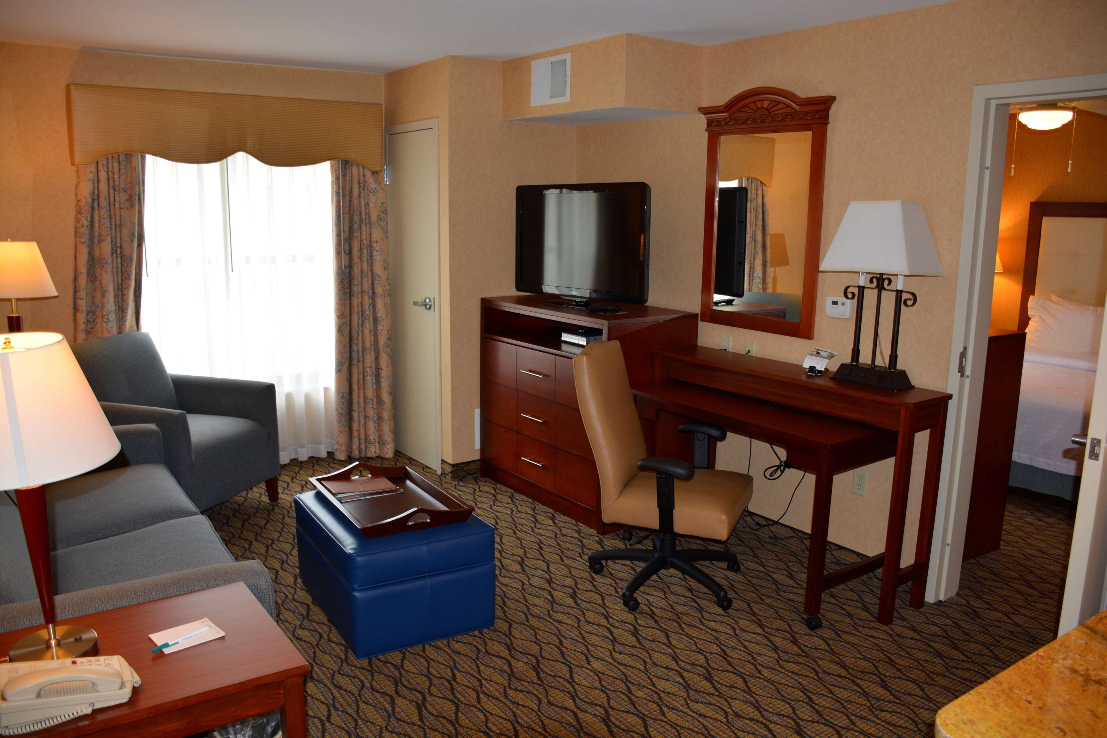 Quarto de hotel com sofá, escrivaninha, computador, televisão, espelho e abajur.