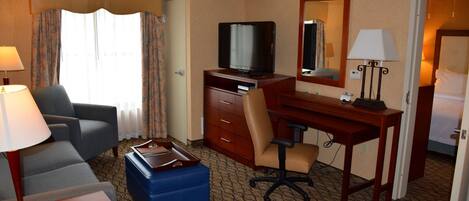 Quarto de hotel com sofá, escrivaninha, computador, televisão, espelho e abajur.