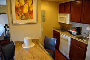 Una pequeña cocina con microondas, cafetera y lavamanos. Hay una silla y una mesa con una cafetera encima.