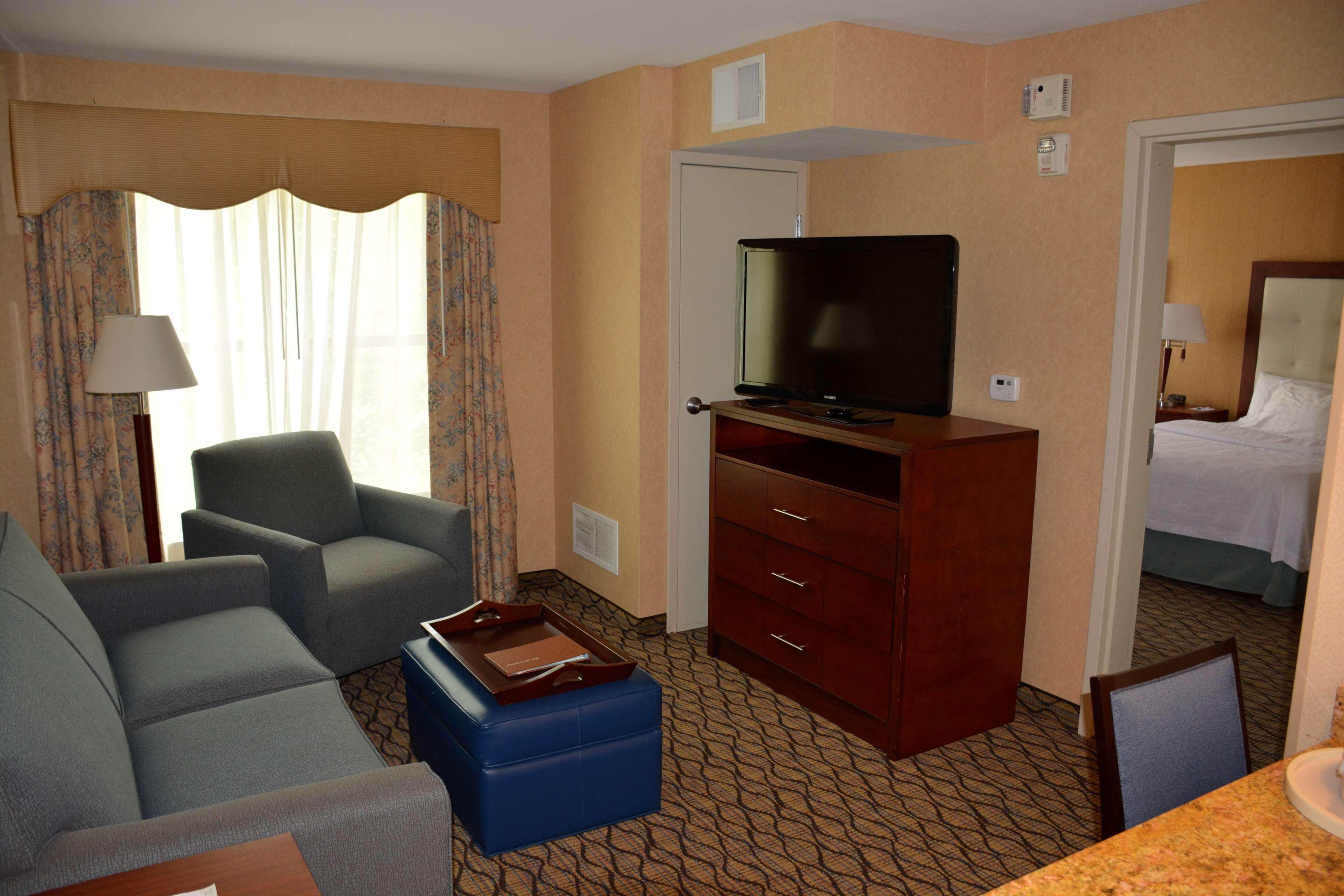 A hotel room with a sofa, a television on a wooden stand, a desk with a lamp, and a bed in the adjacent room.