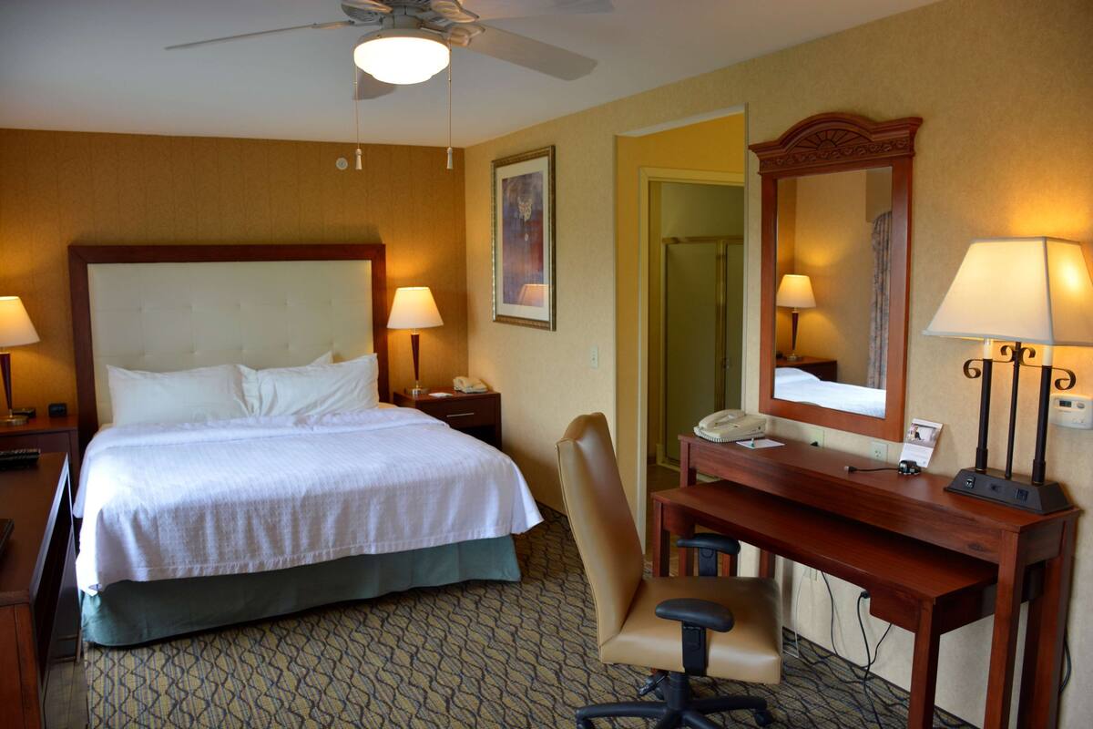 A hotel room with a large bed, a desk with a chair, a desk lamp, a telephone, and a mirror.