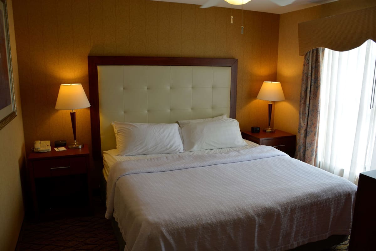 A hotel room with a large bed, two bedside tables with lamps, a nightstand, and a window with curtains.