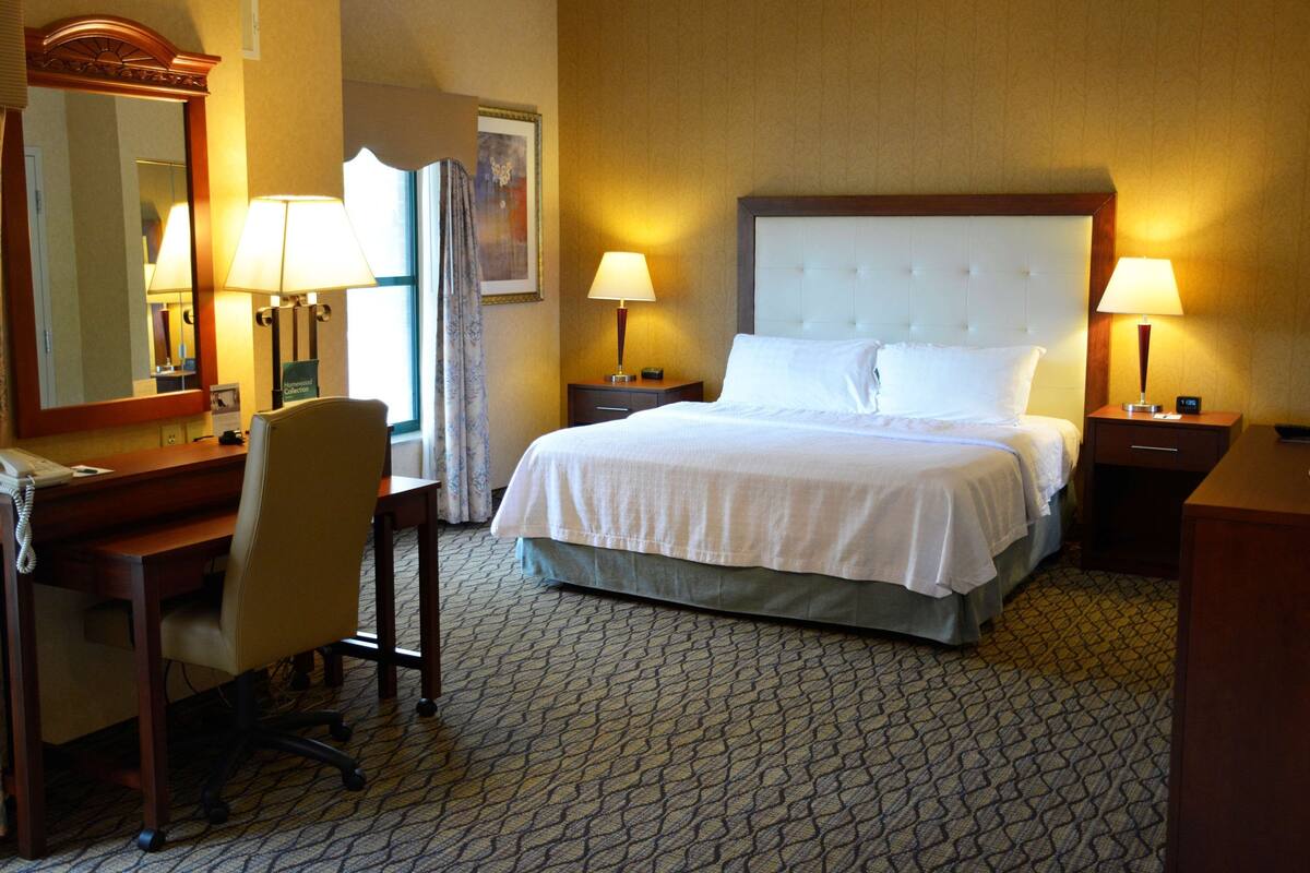 Une chambre d’hôtel avec un grand lit, deux tables de chevet avec des lampes, un bureau avec une chaise, un grand miroir et une moquette à motifs.