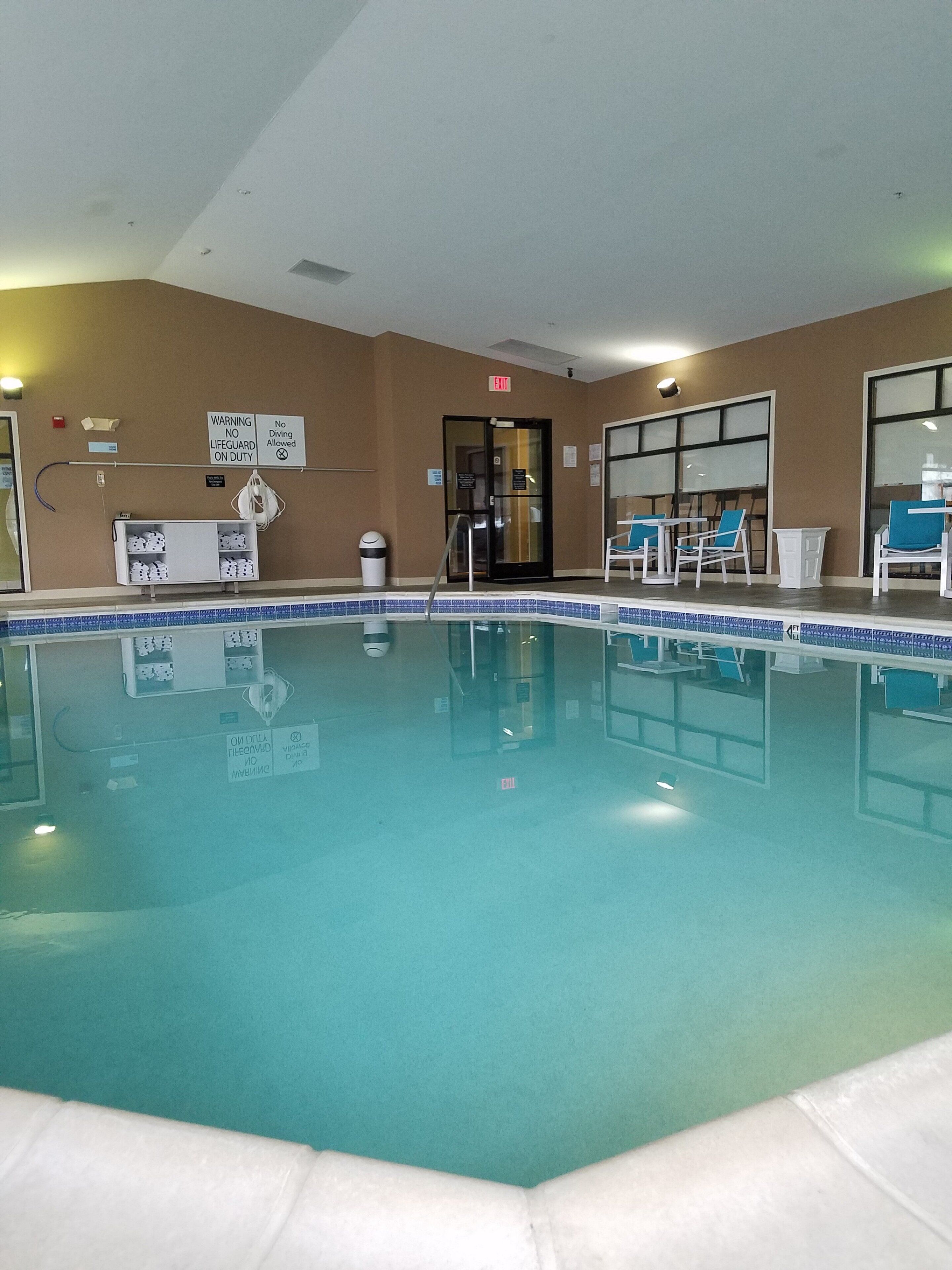 An indoor swimming pool with a lifeguard station, chairs, and a clear exit sign.