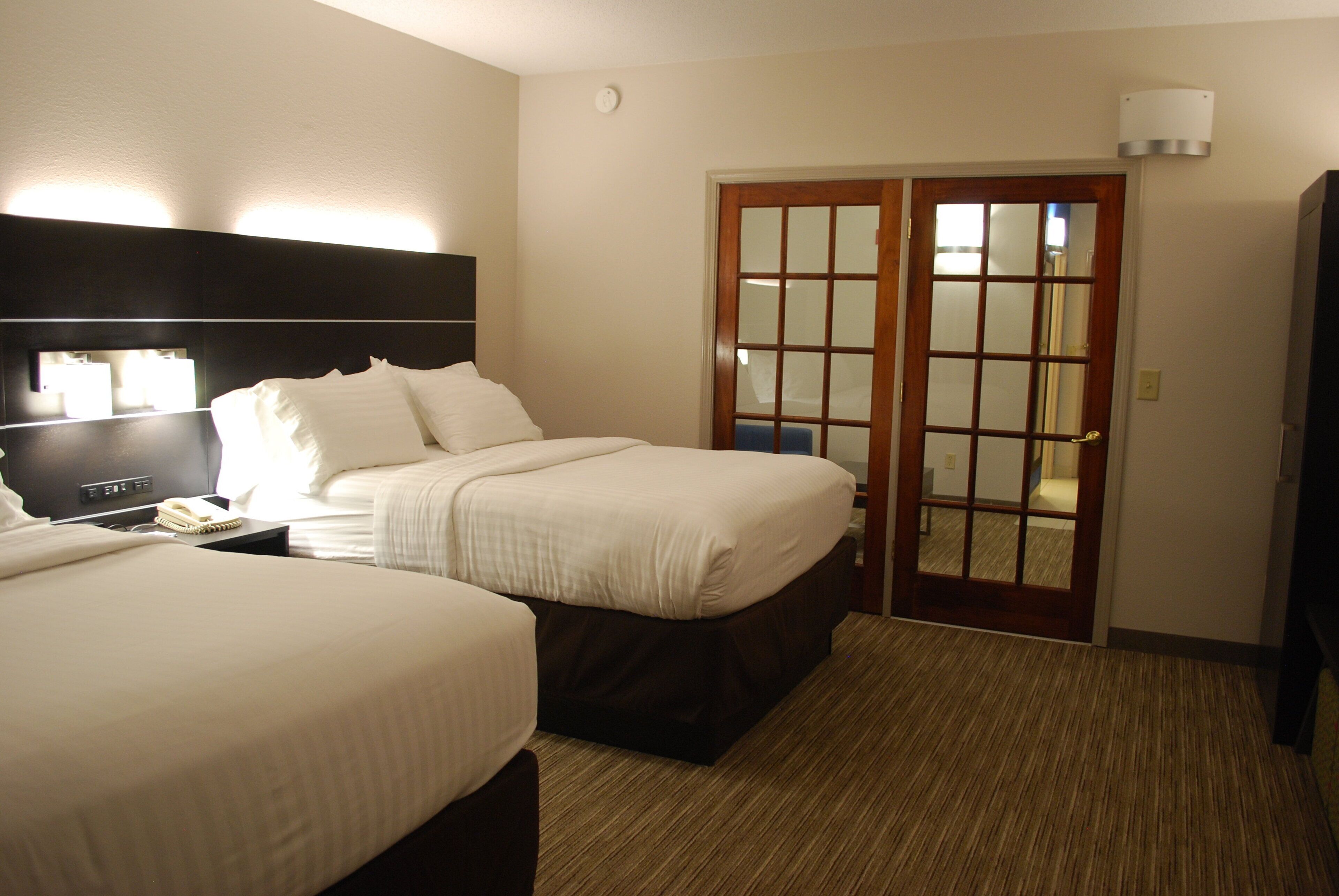 A hotel room with two beds, a bedside table with a lamp, a mirror, and a door with a glass panel.