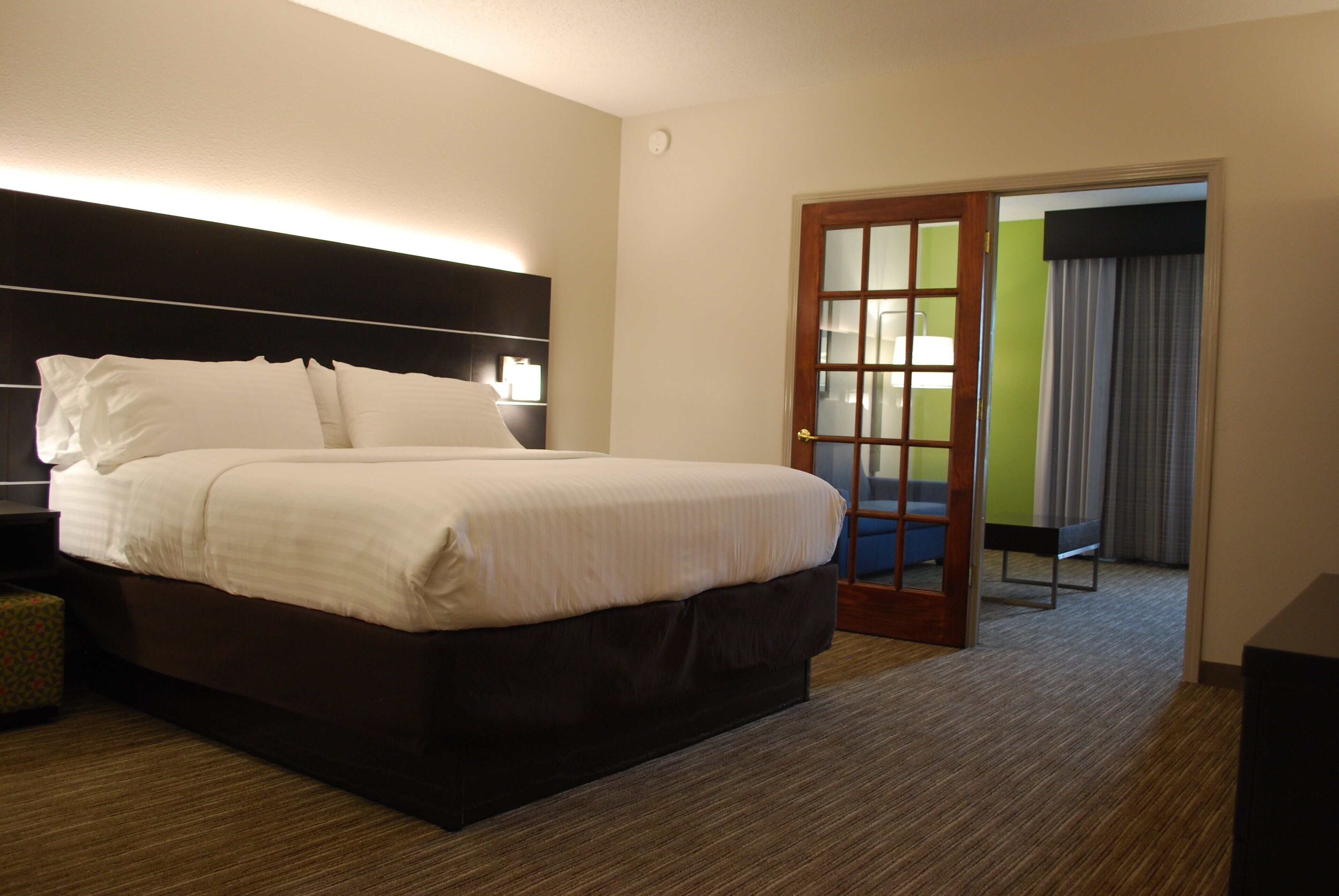 A hotel room with a large bed, a nightstand, a wall-mounted lamp, and a door leading to a bathroom.