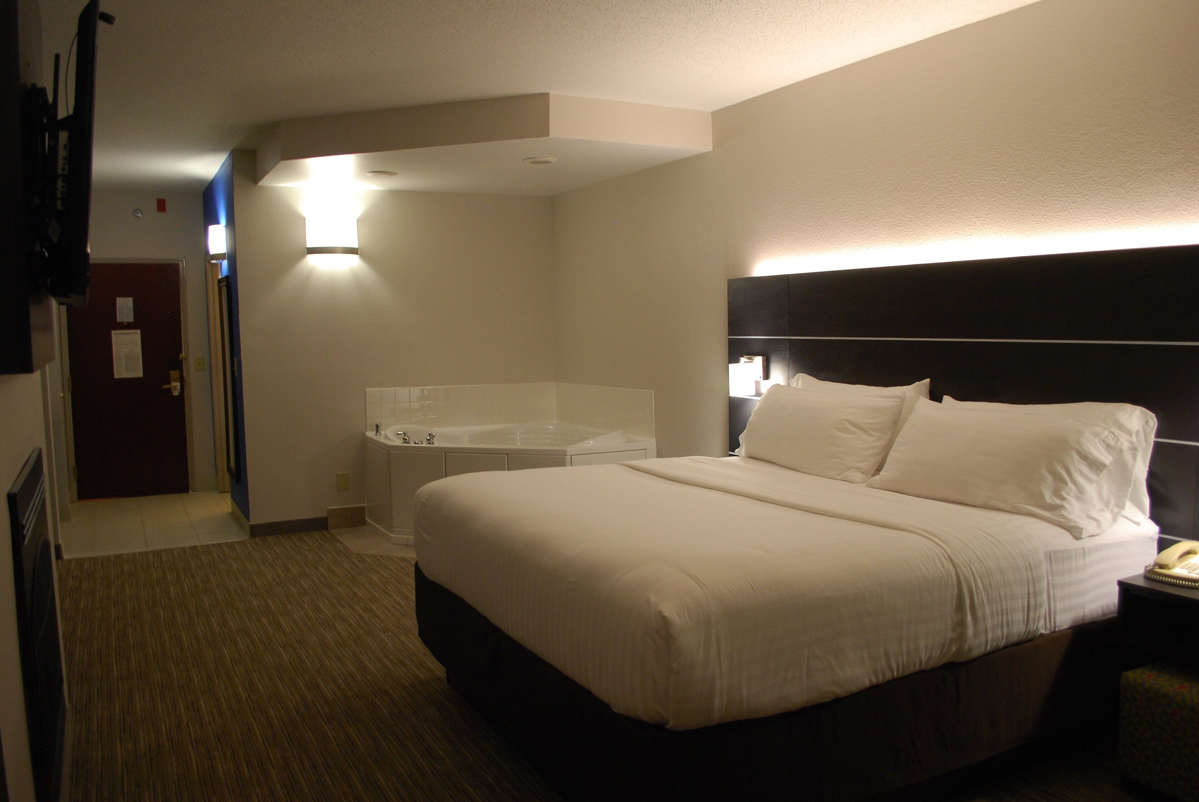 A hotel room with a large bed, a bedside table with a lamp, a bathroom with a bathtub, and a door leading to another room.