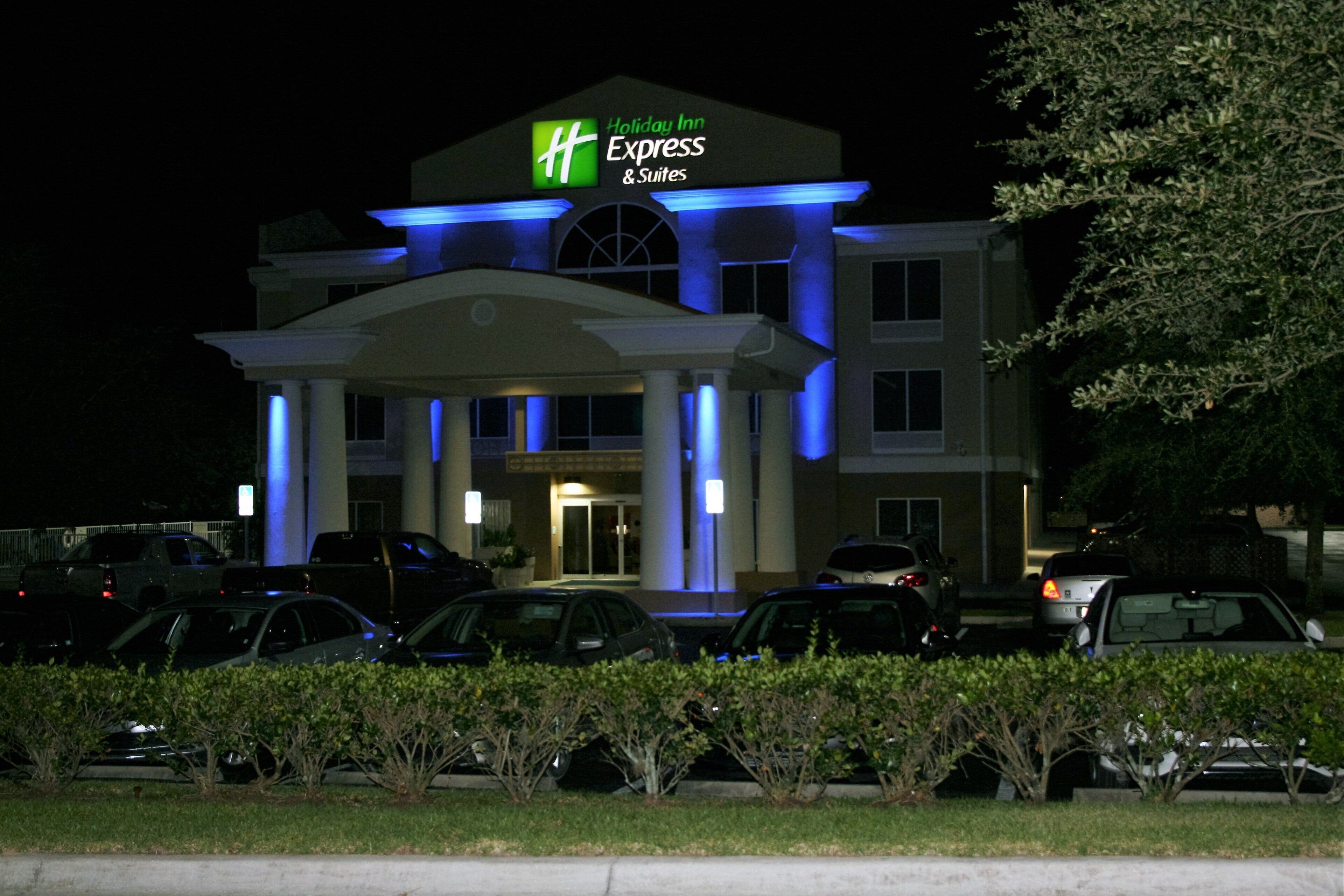 A Holiday Inn Express & Suites building with a central entrance, parking lot, and surrounding greenery.