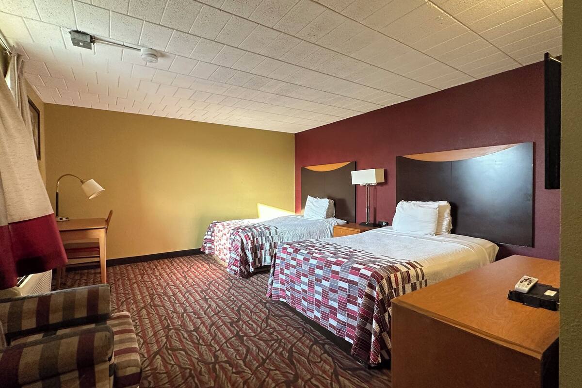 A hotel room with two beds, a desk, a chair, and a lamp.