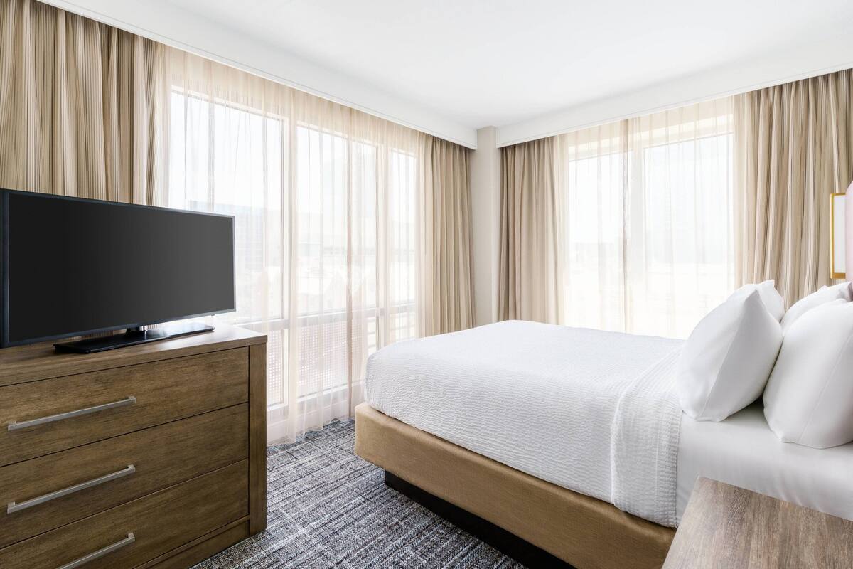 A hotel room with a bed, a TV on a wooden dresser, and sheer curtains.