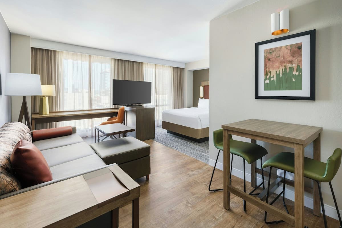 A modern hotel room with a sofa, a dining table with chairs, a bed, a TV, and a painting on the wall.
