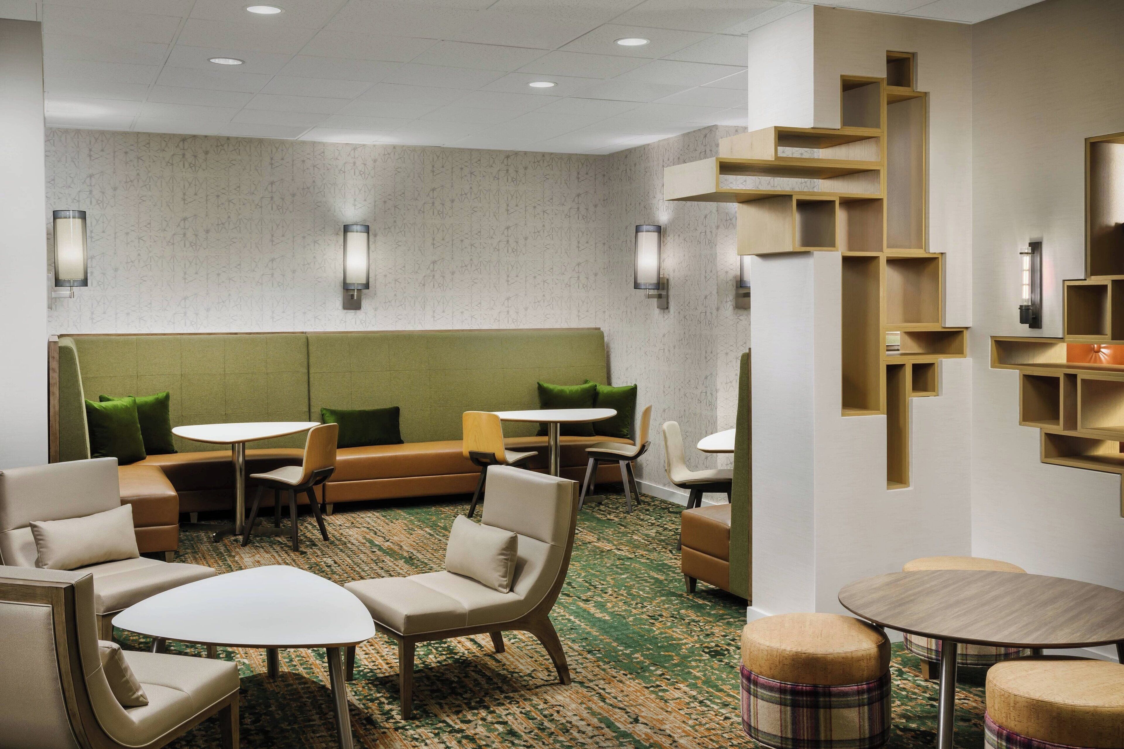 A modern hotel lobby with a green sofa set, multiple seating areas, and a unique wooden shelving unit.