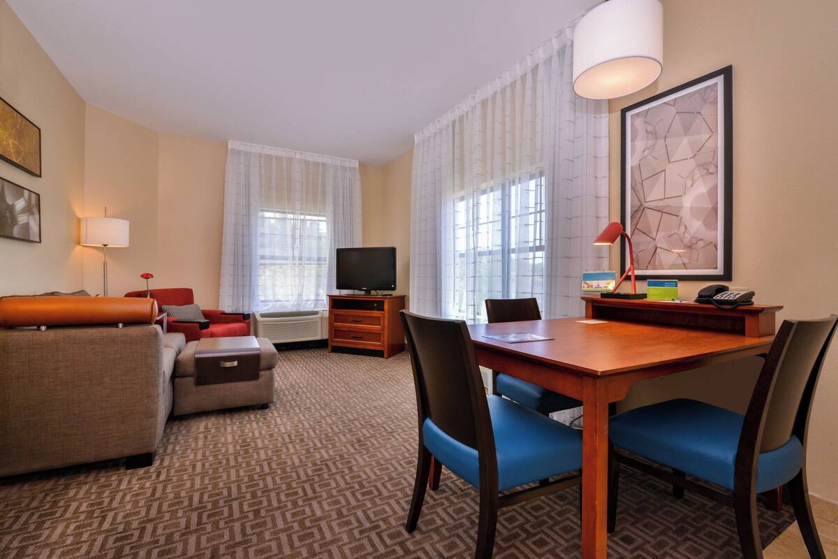 Habitación de hotel con sofá, una mesa de comedor con sillas, televisor, lámpara y un cuadro en la pared.
