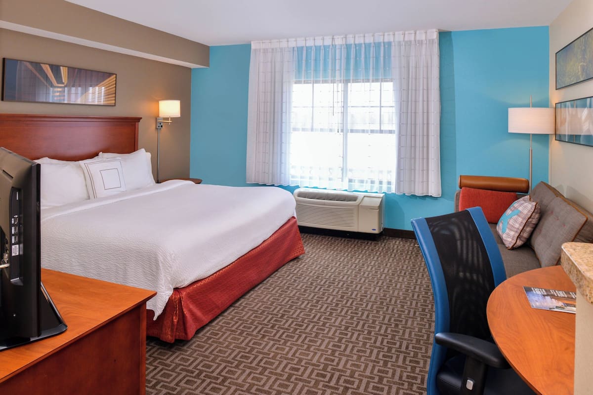 Habitación de hotel con una cama grande, un escritorio con computadora, una silla, una mesa, un ventanal con cortinas y un televisor de pared.