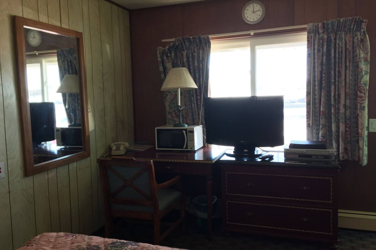 A hotel room with a wooden desk, a television, a lamp, a chair, a dresser, and a bed with a floral bedspread.