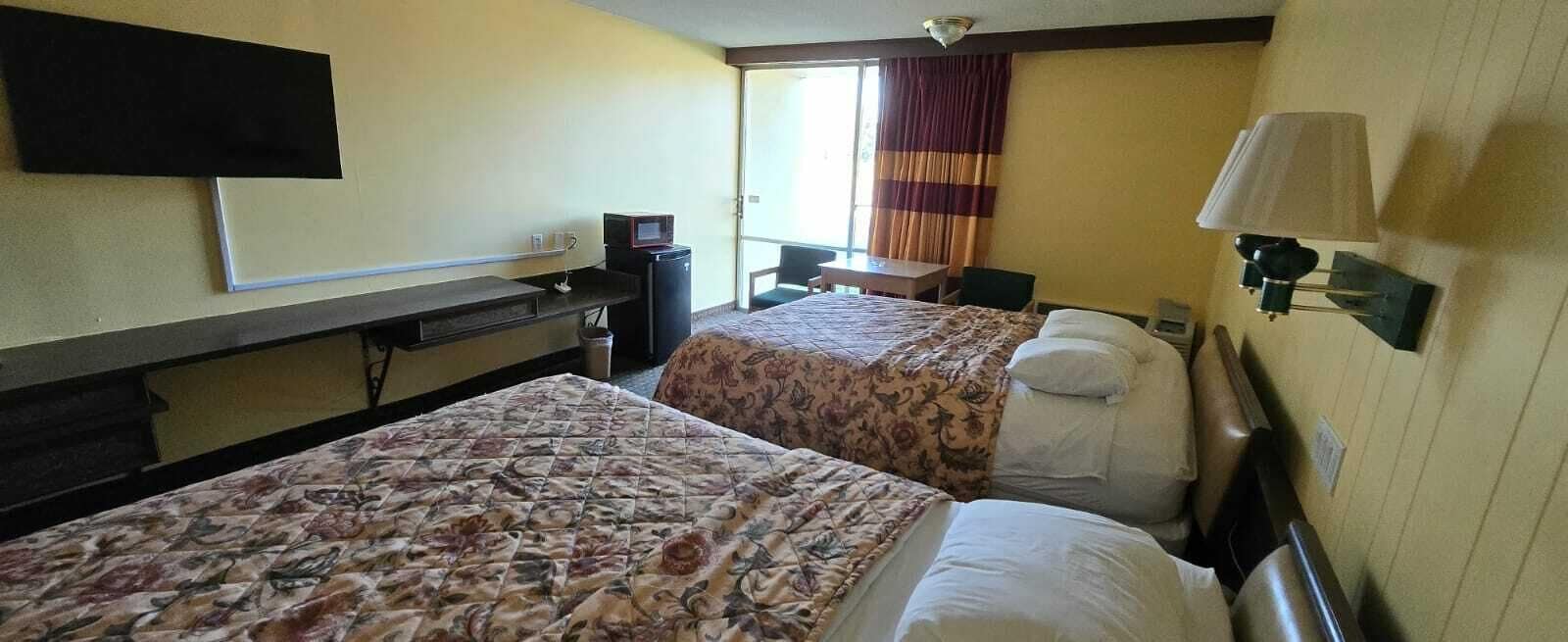 Una habitación de hotel con dos camas, un escritorio con computadora, un televisor y una ventana con cortinas.