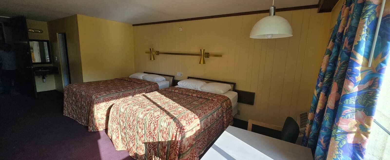 A hotel room with two beds, a desk, a chair, a hanging lamp, and a window with curtains.