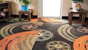 Una habitación con alfombra estampada, dos plantas en macetas, un estante de madera con libros y una puerta con un cartel.