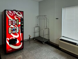 Máquina de vendas com diversas opções de bebidas, um carrinho de metal e uma janela com persianas.