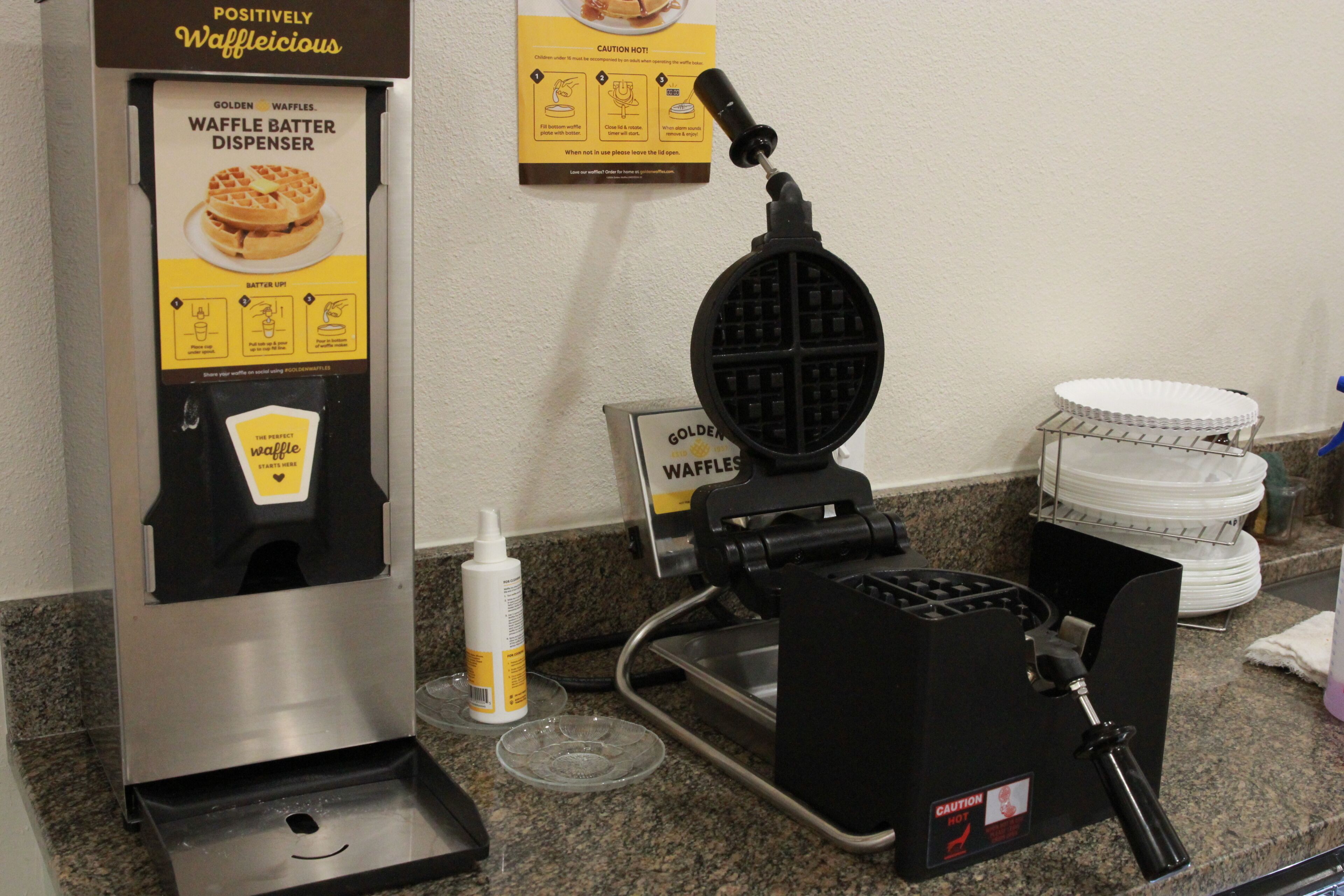 A waffle batter dispenser with instructions, a waffle maker, and a stack of paper plates on a counter.