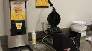 A waffle batter dispenser with instructions, a waffle maker, and a stack of paper plates on a counter.