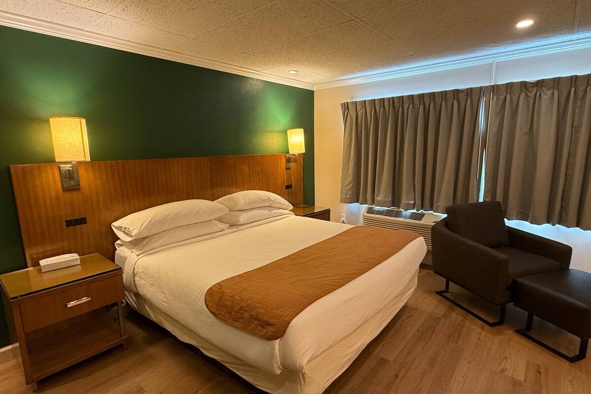 A hotel room with a bed, bedside tables, a chair, a window with curtains, and a wall-mounted air conditioning unit.