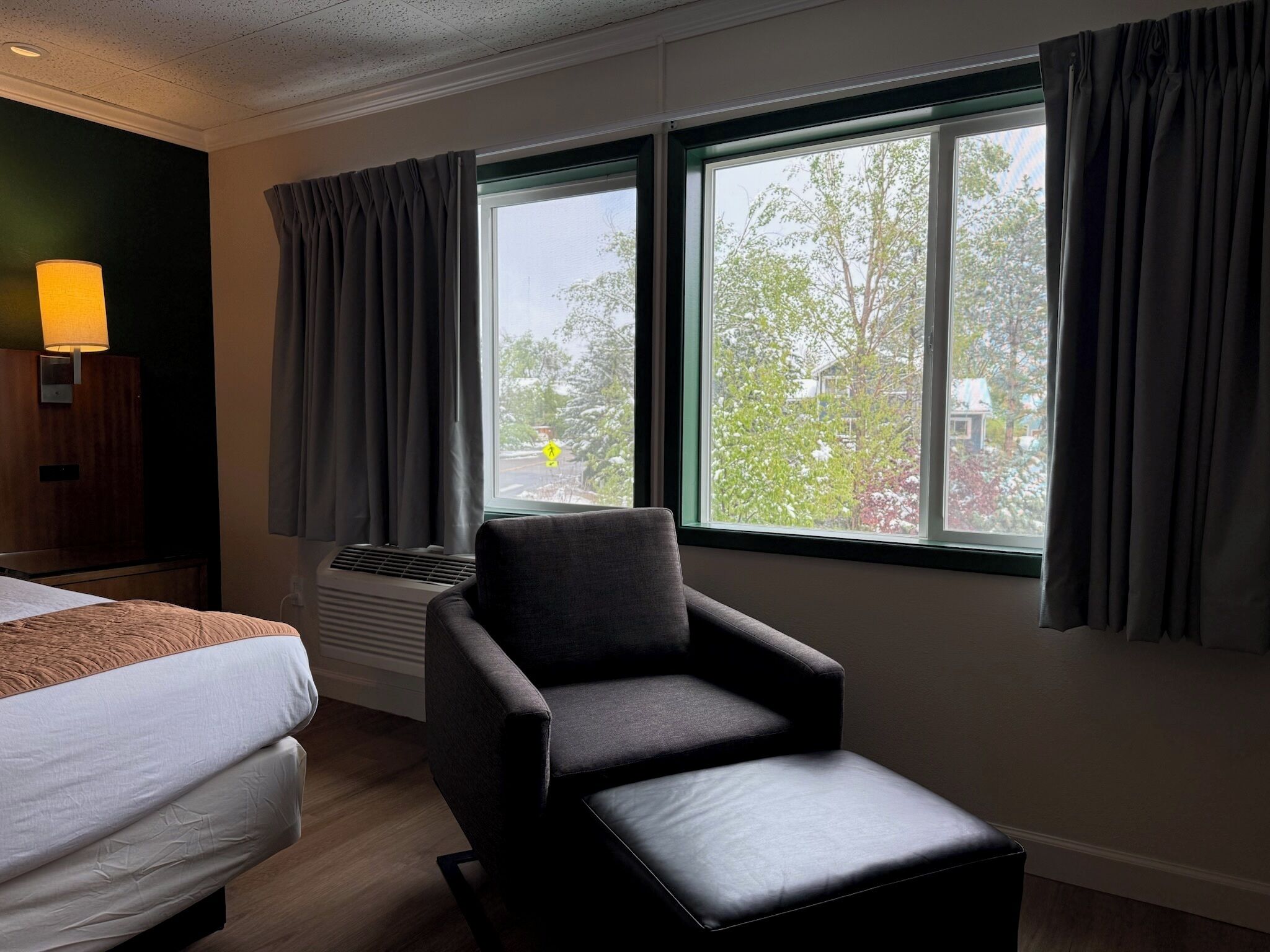 Habitación de hotel con cama, una silla, ventana con cortinas, lámpara de pared y radiador.