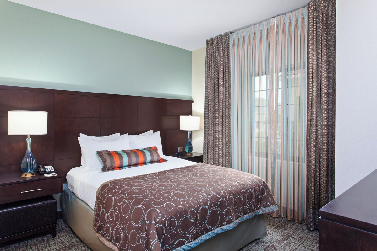 A hotel room with a large bed, two bedside tables with lamps, a nightstand, a dresser, and a window with curtains.