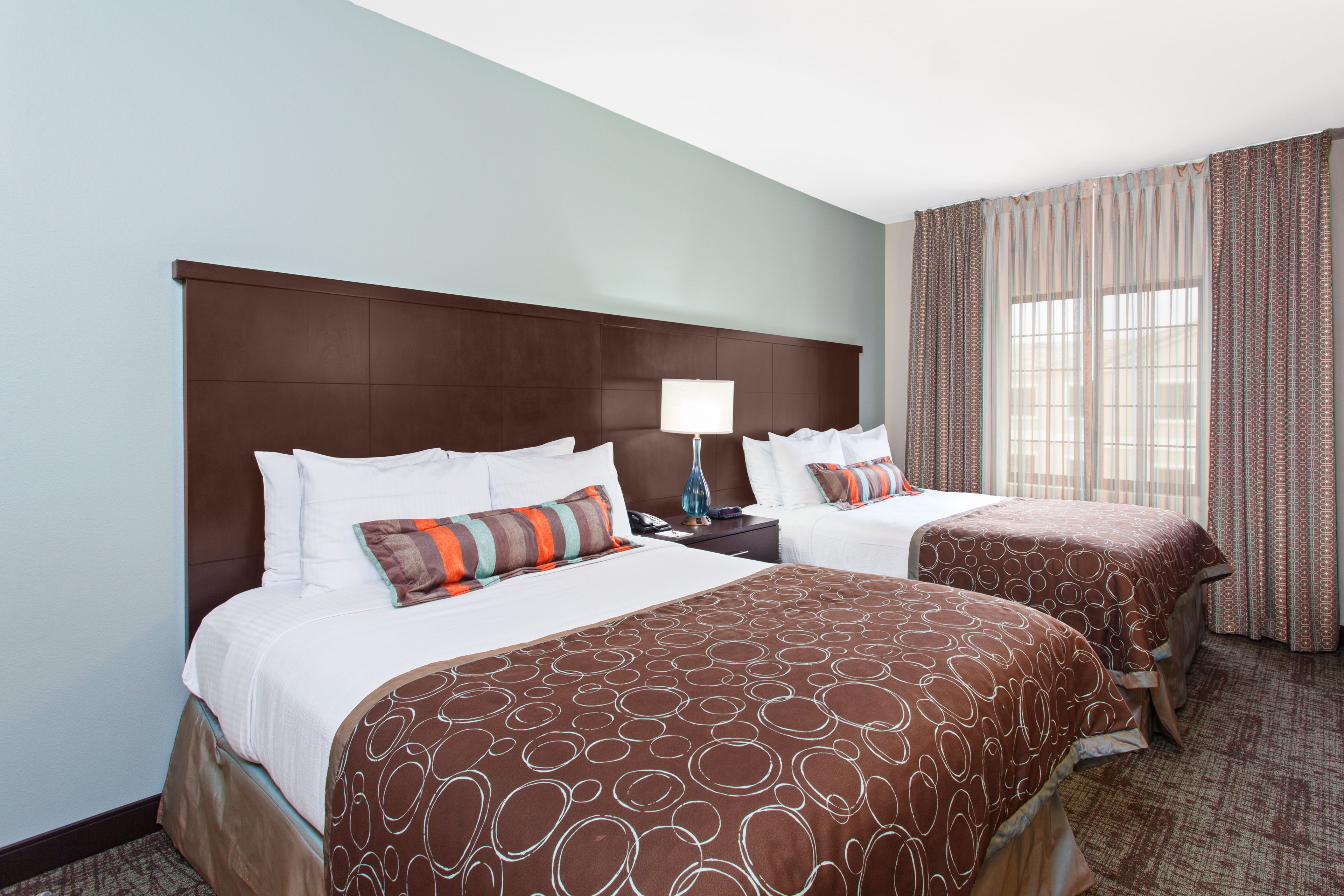 Quarto de hotel com duas camas, uma cabeceira grande, uma mesa de cabeceira com abajur e uma janela com cortinas.