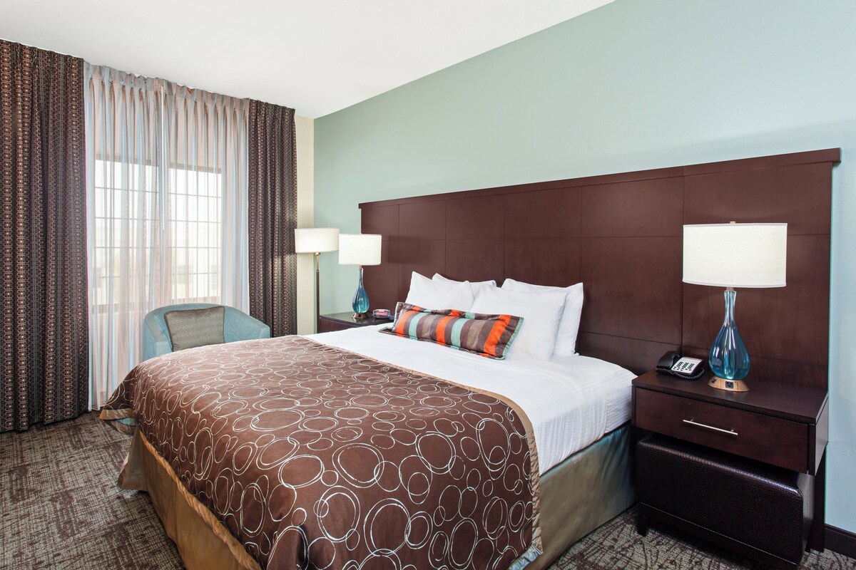 A hotel room with a large bed, two bedside tables with lamps, a chair, and a window with curtains.