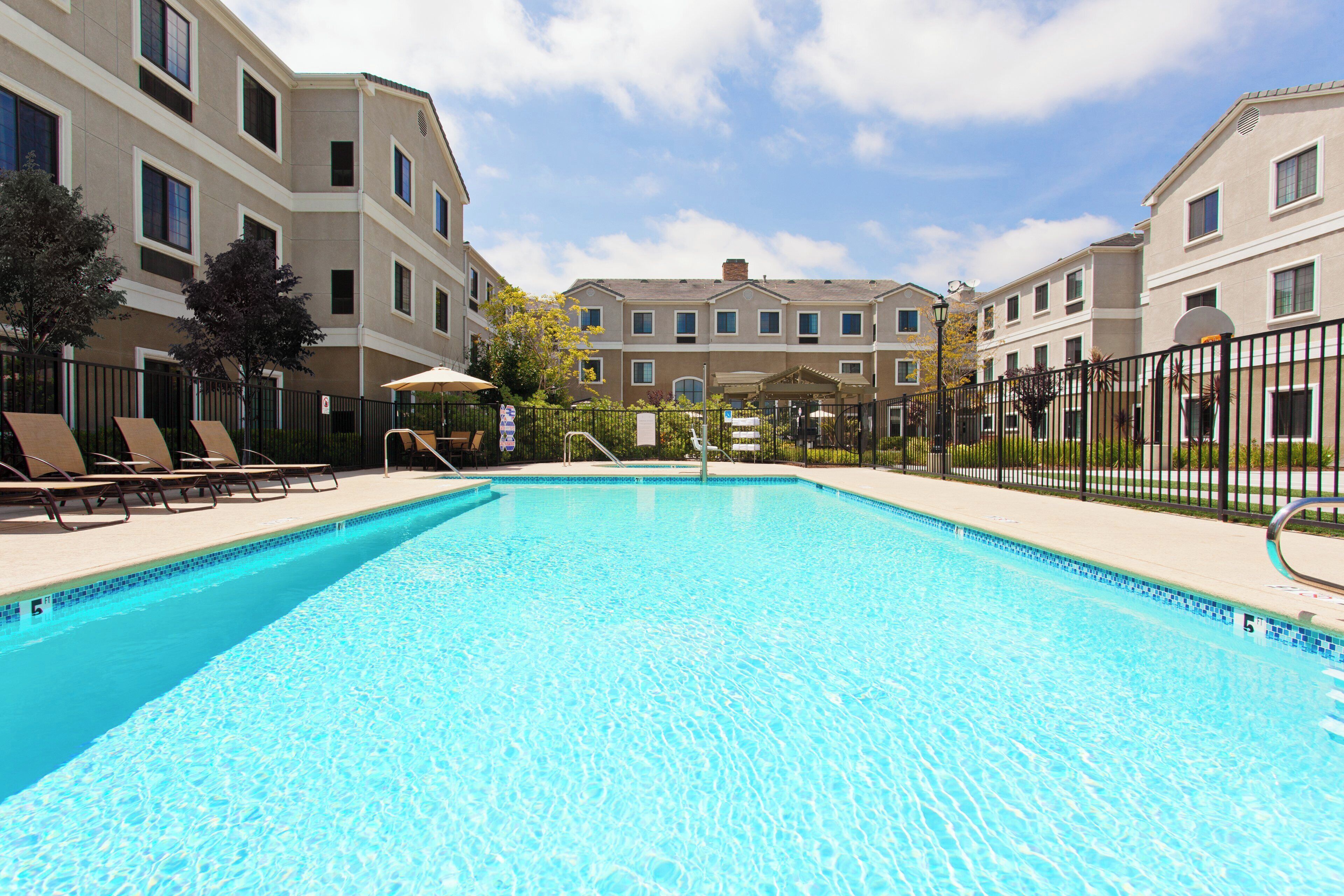 A swimming pool with lounge chairs and a covered seating area, surrounded by a fence and buildings in the background.