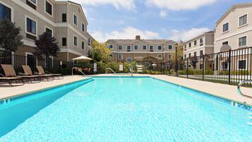 A swimming pool with lounge chairs and a covered seating area, surrounded by a fence and buildings in the background.