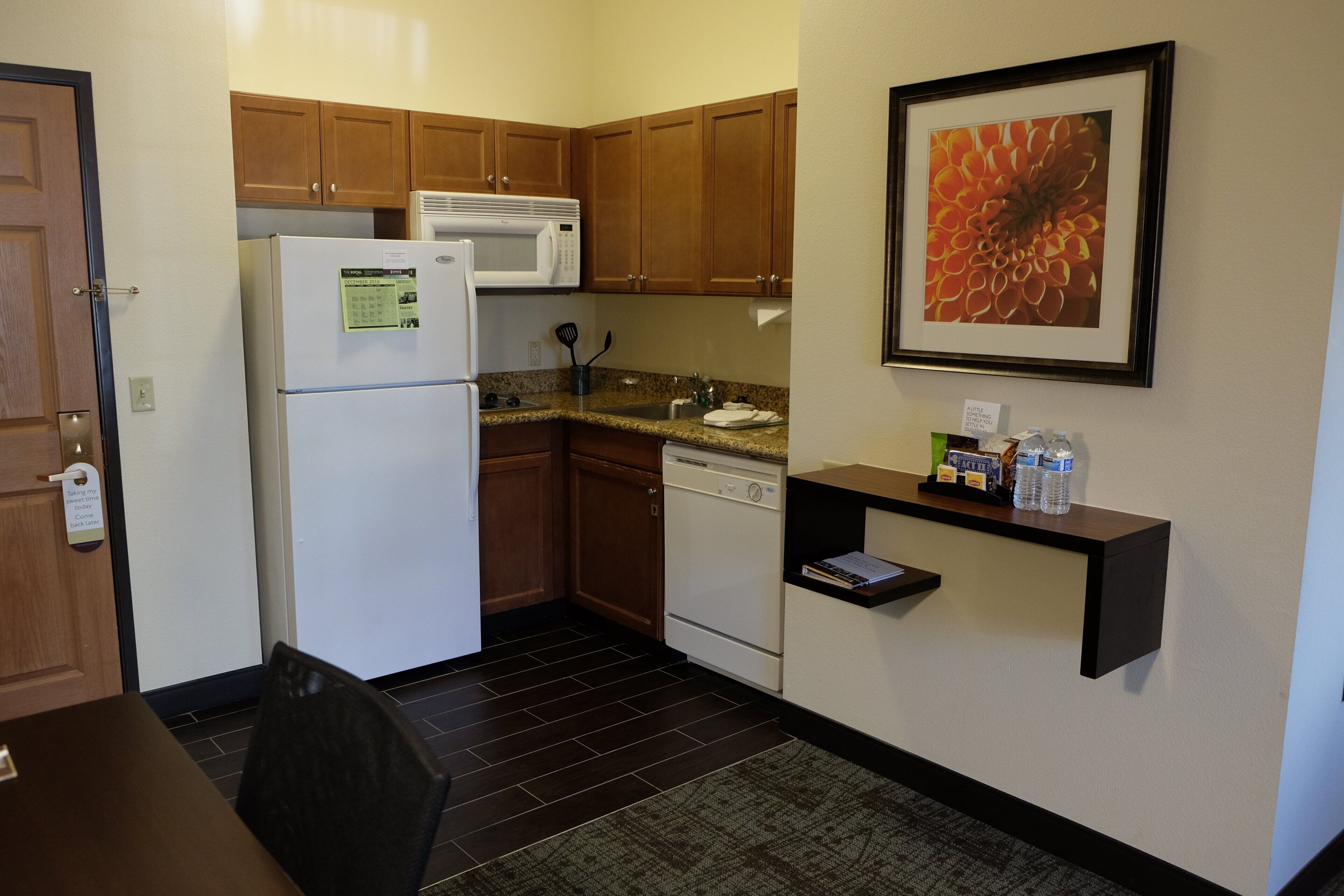 A compact kitchen with a refrigerator, microwave, and dishwasher. There is a small shelf with bottled water and a framed picture on the wall.