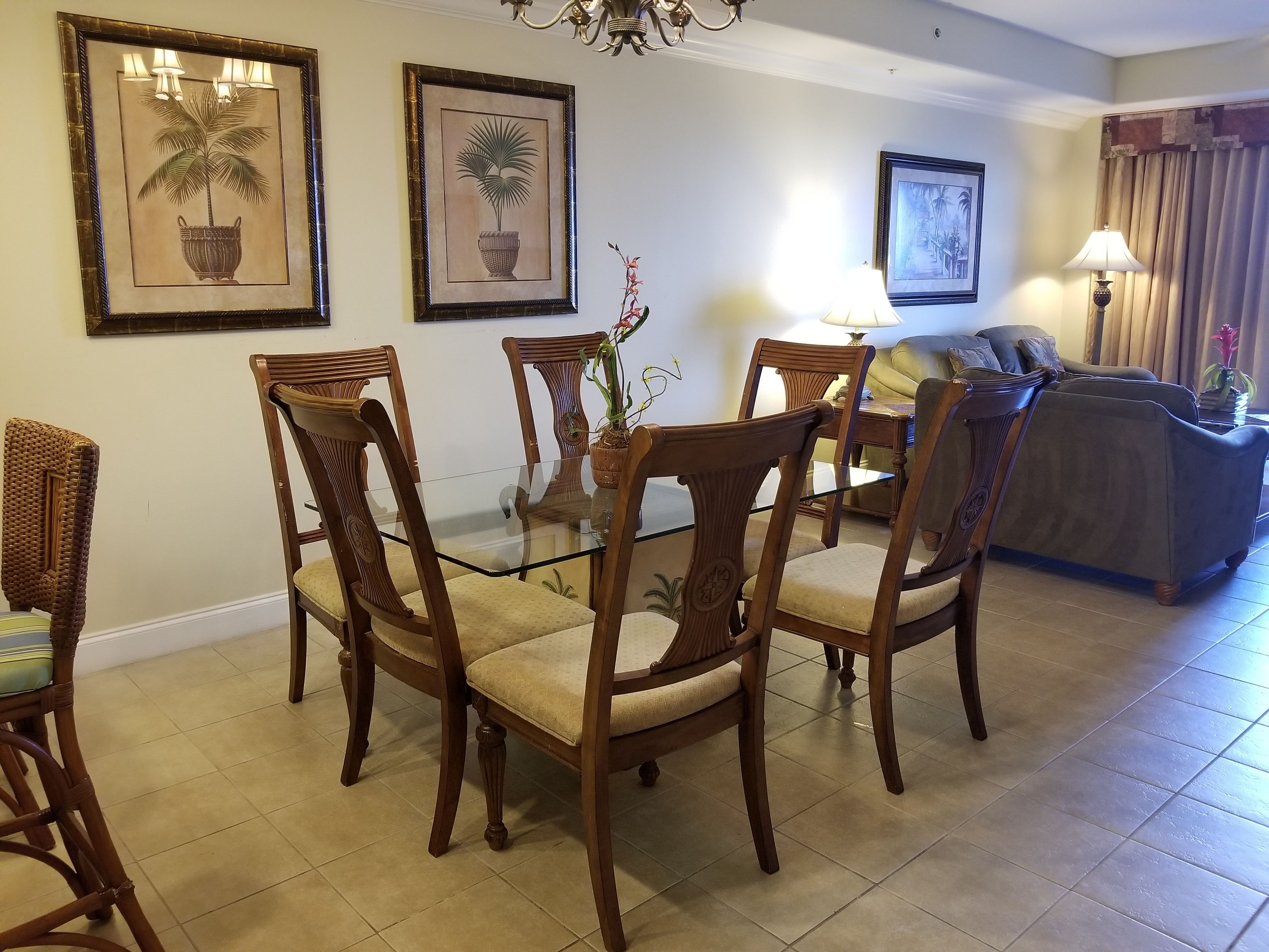 Uma área de refeições com mesa de vidro rodeada por cadeiras de madeira, um sofá e quadros nas paredes.