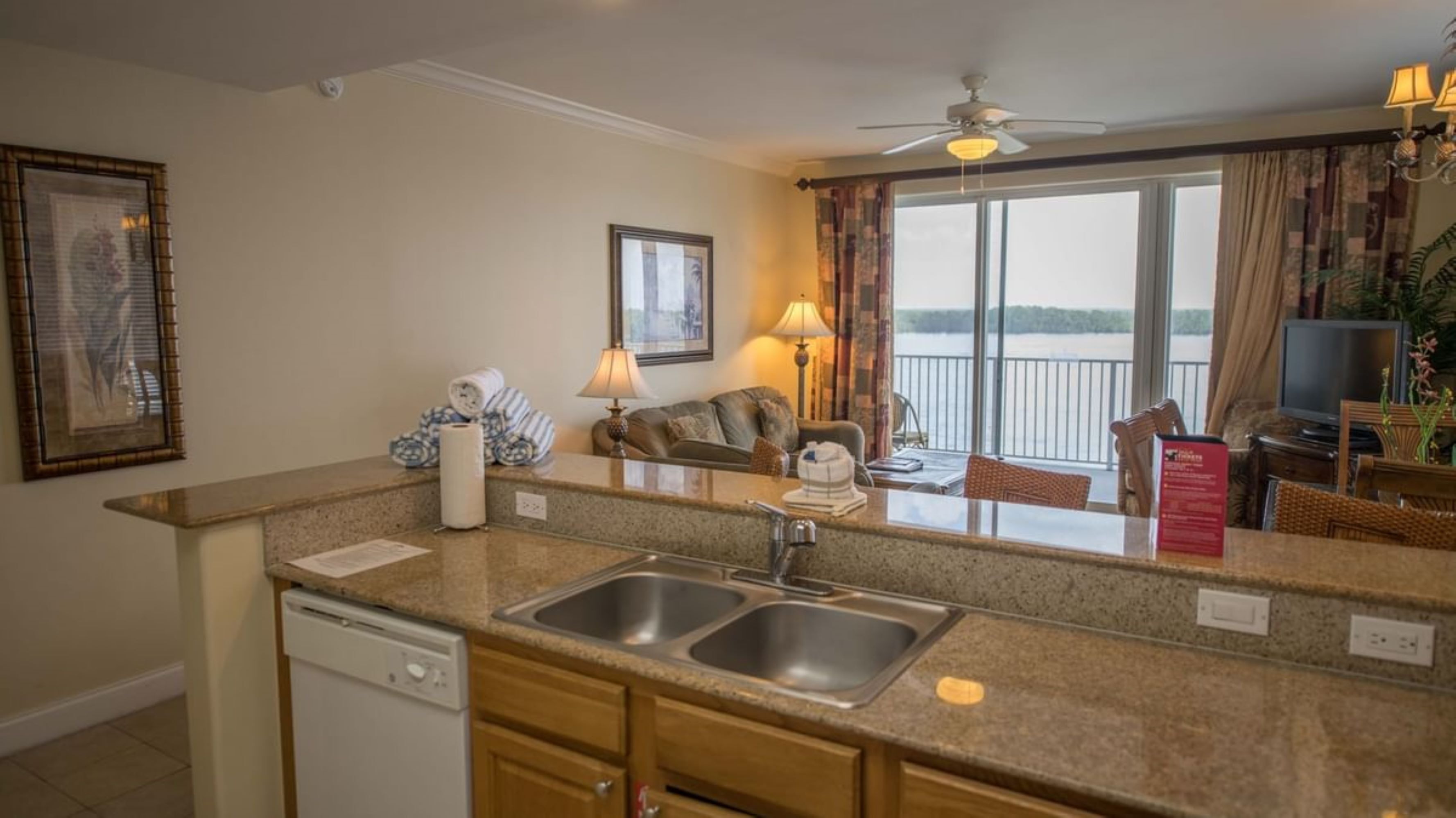 A kitchen with a dishwasher, double sink, and a dining area with a table and chairs. There is a balcony with a view of the water.