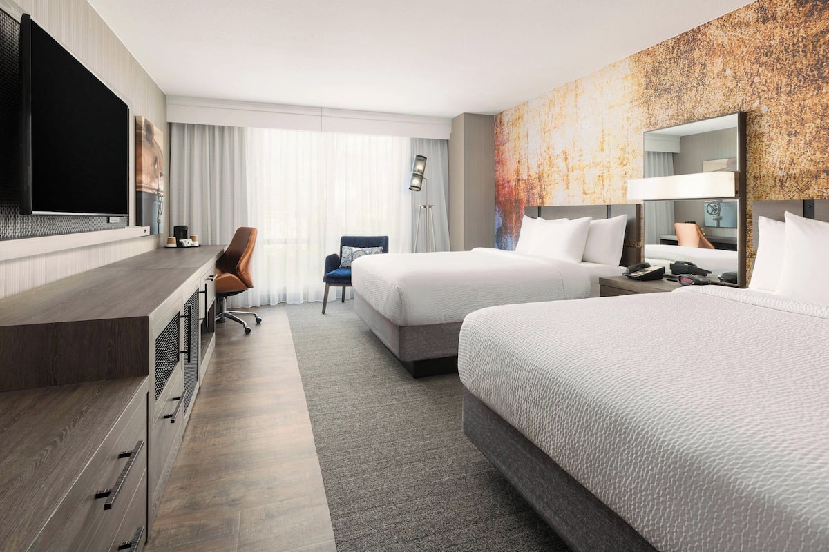A hotel room with two beds, a desk with a flat-screen TV, a chair, and a large mirror.