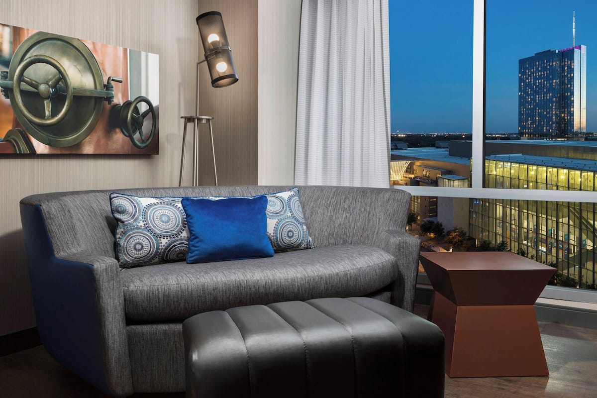 A modern living room with a grey sofa, blue and patterned cushions, a black ottoman, a floor lamp, and a large window with a city view.