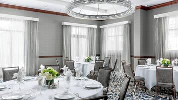 En hotels restaurant med runde borde dækket til et måltid, store vinduer med gardiner og en lysekrone.