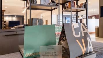 A hotel reception area with a 'sweet' branded bag, a 'CA' branded bag, and a sign instructing to 'collect your delivery order here.'