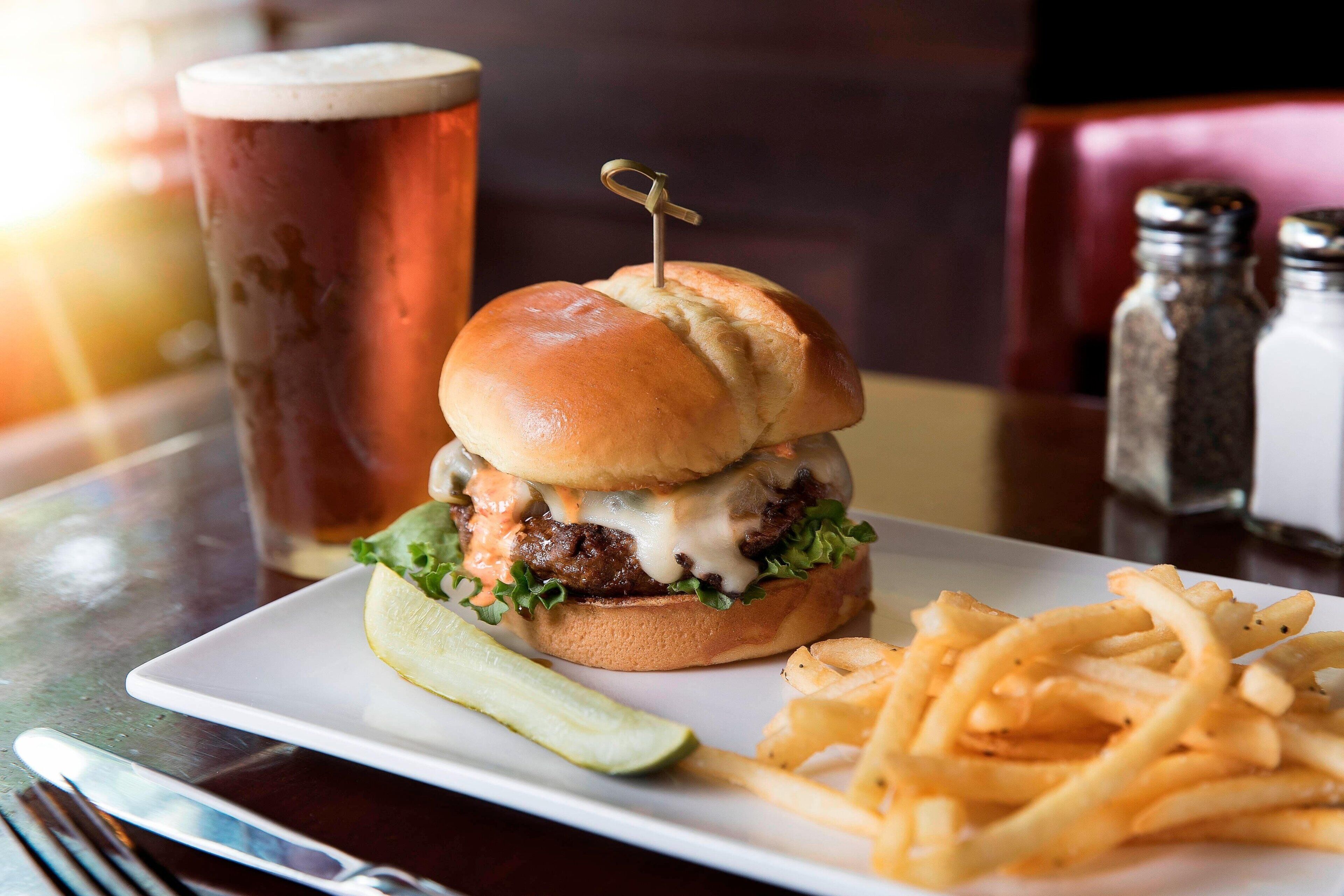 Ein Burger mit Gurke und Pommes auf einem Teller, ein Glas Bier und Salz- sowie Pfeffermühlen auf einem Tisch.