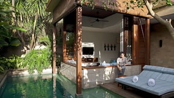 Relax by the serene pool surrounded by lush greenery and a wooden pergola, with a lounge chair and bar area nearby.