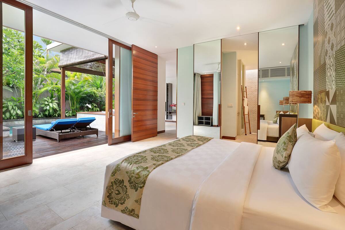 Quarto moderno com uma cama grande, porta de vidro que dá para uma varanda com espreguiçadeira e vista para vegetação exuberante.